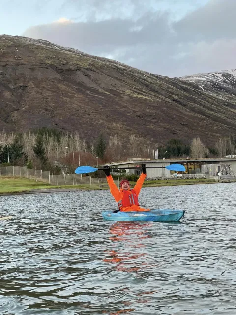Viktor orri úti á vatni í kajak. Hann heldur árinni fyrir ofan höfuð eins og hann sé að fagna. í bakgrunni er fjall og íslensk náttúra.